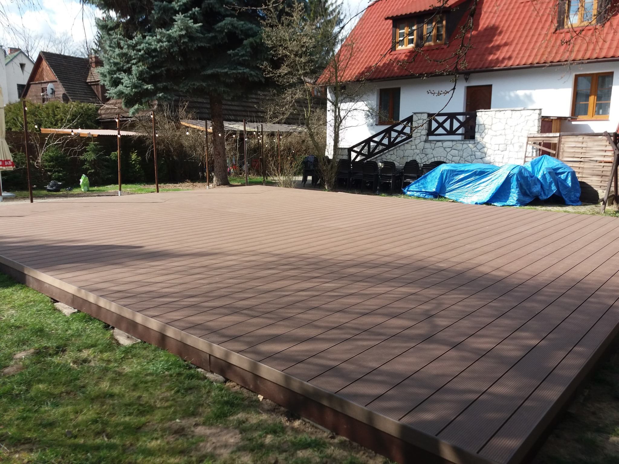 Rozległy, brązowy taras z desek kompozytowych na trawniku, w tle dom z czerwoną dachówką i zabudowa z pergolą.