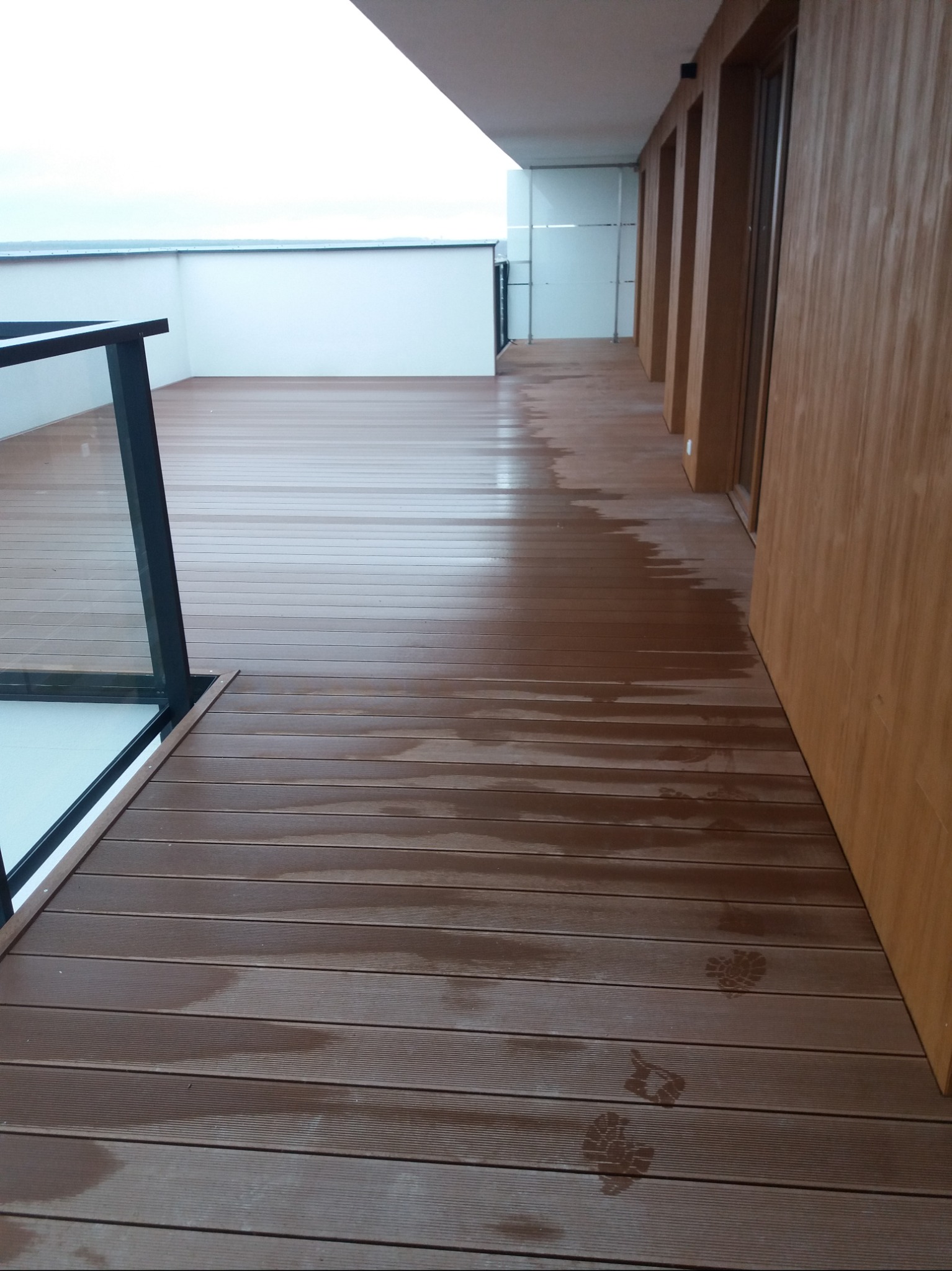 Brązowy taras z desek kompozytowych, częściowo mokry, z widocznymi odciskami butów, z czarną balustradą i drewnianą ścianą budynku w tle.