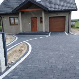 Nowoczesny podjazd z szarej kostki brukowej z białym obramowaniem, prowadzący do domu z brązową bramą garażową i drewnianymi drzwiami wejściowymi; widoczny fragment przygotowanej rabaty.