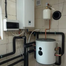 Biały piec centralnego ogrzewania marki Rotenso z czarnymi rurami w kotłowni wyłożonej kafelkami.