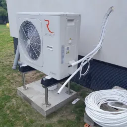 Zewnętrzna jednostka pompy ciepła Rotenso Aquami Series zamontowana na betonowym fundamencie z metalowymi wspornikami, widoczne białe rury instalacyjne prowadzące do otworu w ścianie budynku.