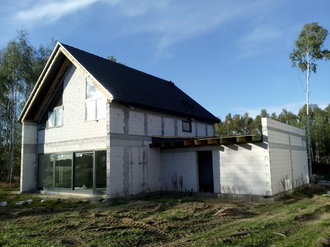 Surowy stan domu jednorodzinnego w trakcie budowy, widoczne białe bloczki, czarny dach, duże okna i przygotowane zadaszenie nad wejściem, otoczenie stanowią drzewa i niezagospodarowany teren.