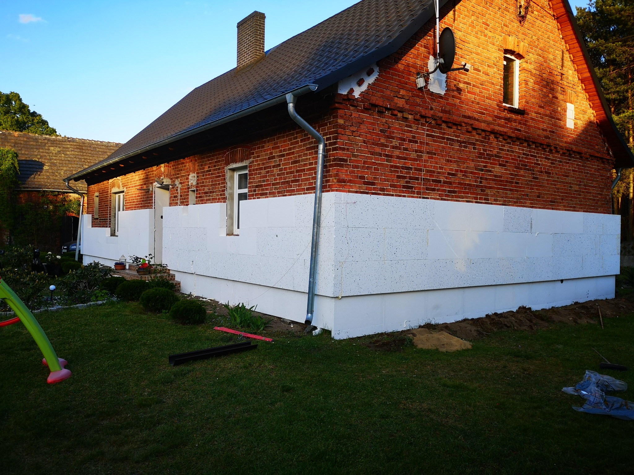 Częściowo ocieplony dom z czerwonej cegły, widoczne białe płyty izolacyjne na dolnej części elewacji, zieleń trawnika i elementy placu zabaw w kadrze.