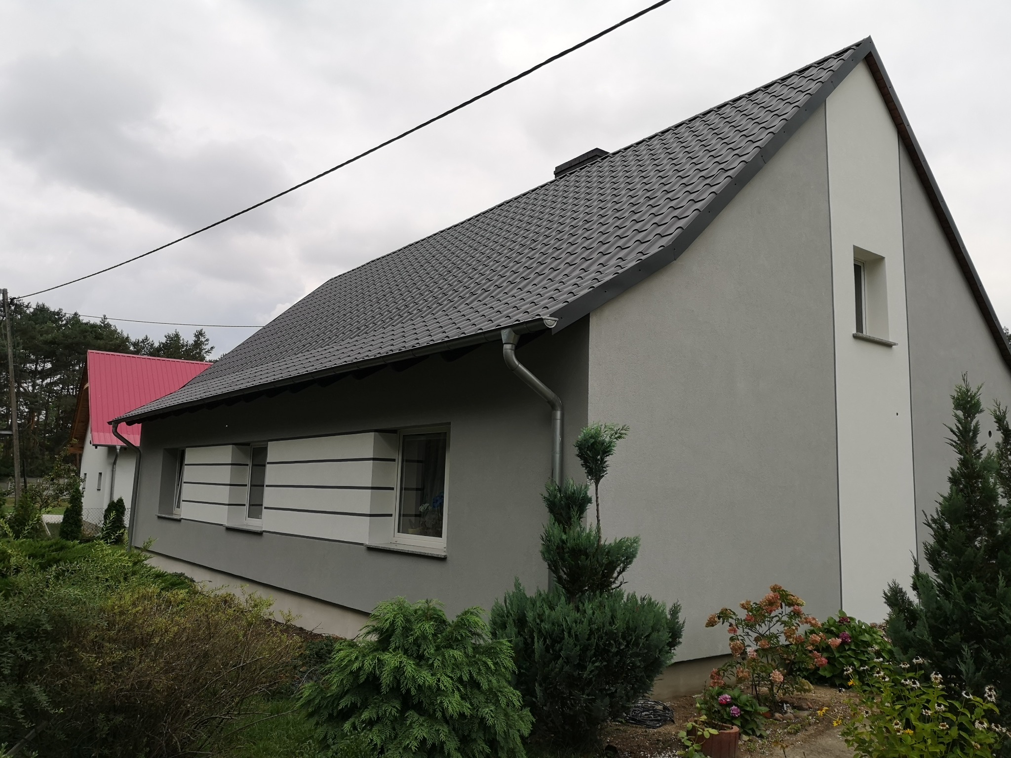 Nowoczesny dom jednorodzinny z szarą elewacją, grafitowym dachem i białymi akcentami, otoczony zielenią.