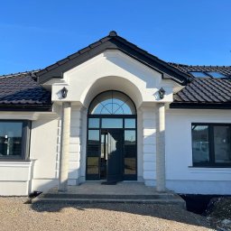 Vladyslav Fedortsov Alternatywa - Elegancki, parterowy dom z białymi ścianami, czarnym dachem i stolarką okienną. Wejście z kolumnami i łukowym oknem. Widoczne detale wykończenia elewacji.