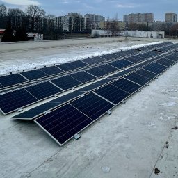 Vladyslav Fedortsov Alternatywa - Rząd paneli fotowoltaicznych na płaskim dachu budynku, częściowo pokrytym śniegiem, z widokiem na zabudowę miejską w tle, pochmurny dzień.