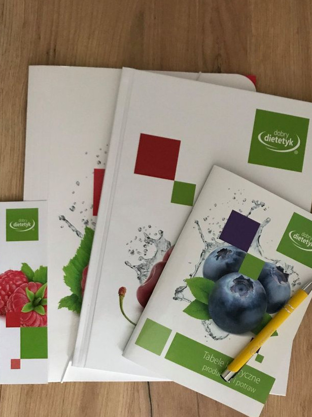 Białe broszury z logo i ilustracjami owoców (maliny, wiśnie, borówki) oraz żółty długopis na drewnianym tle.