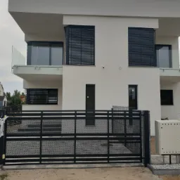 Nowoczesny, dwupiętrowy dom z białymi ścianami, czarnymi żaluzjami zewnętrznymi, szklaną balustradą balkonu i czarną, metalową bramą wjazdową.