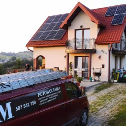 Czerwony samoch&oacute;d dostawczy z napisem 'Fotowoltaika' i numerem telefonu zaparkowany przed domem z panelami słonecznymi na dachu, w tle zielony krajobraz.