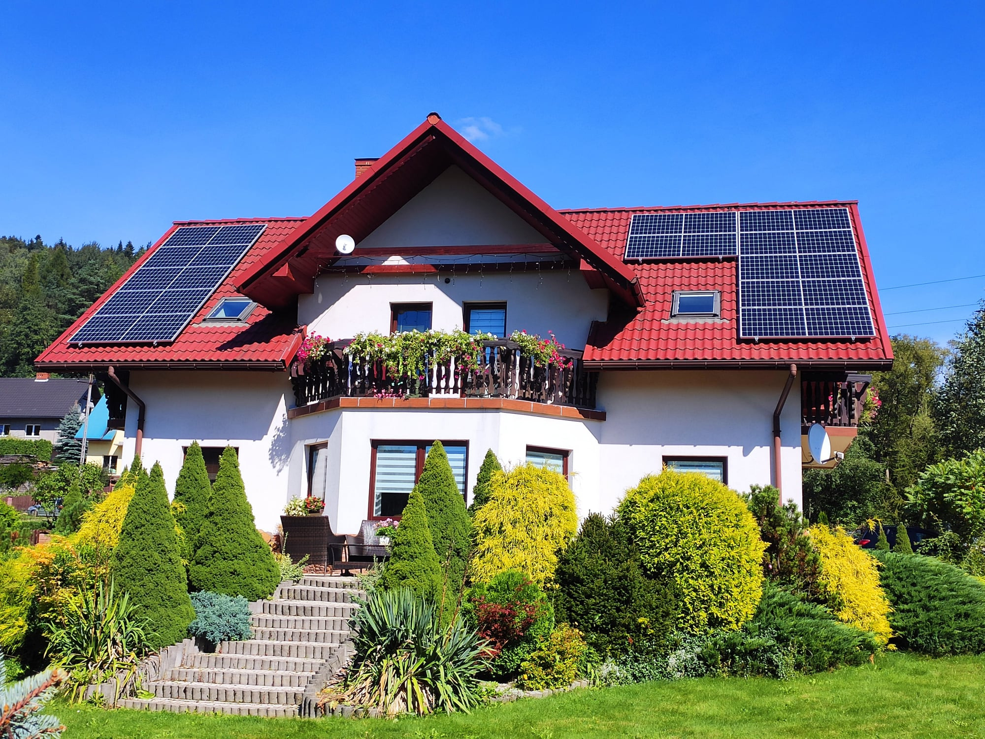 Dom jednorodzinny z czerwoną dachówką i panelami fotowoltaicznymi na dachu, otoczony zielonym ogrodem z ozdobnymi krzewami i schodami wejściowymi.