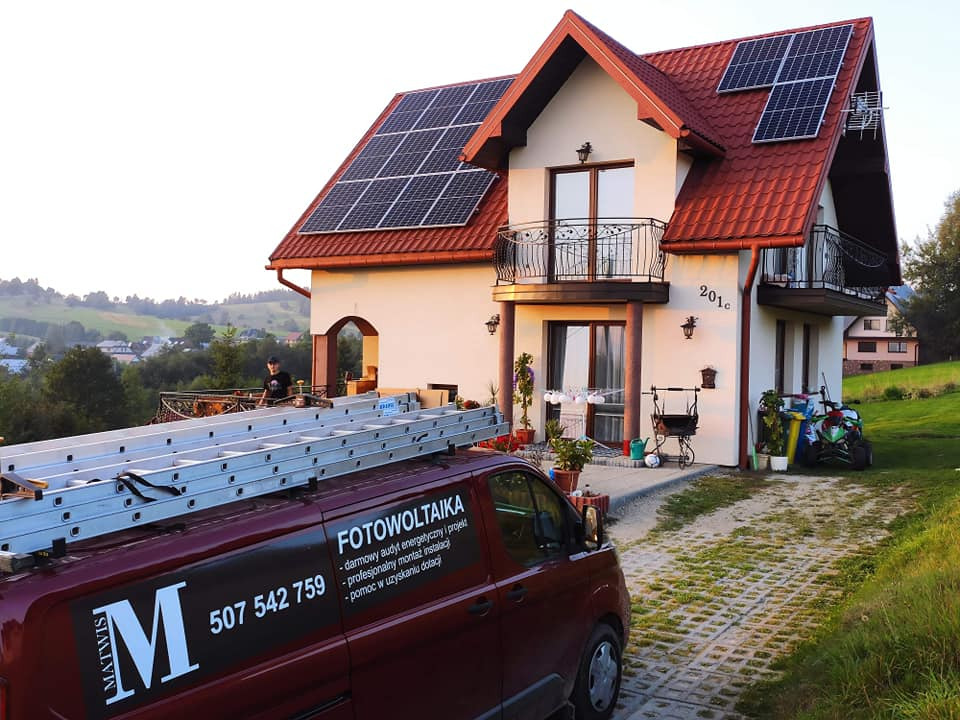 Czerwony samochód dostawczy z napisem 'Fotowoltaika' i numerem telefonu zaparkowany przed domem z panelami słonecznymi na dachu, w tle zielony krajobraz.