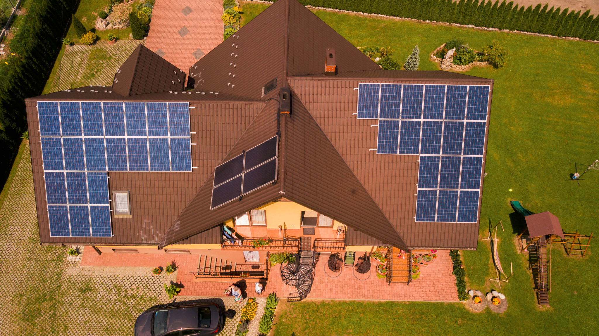 Widok z lotu ptaka na dom z brązowym dachem, na którym zainstalowano panele słoneczne. Dwa zestawy paneli na jednym zboczu dachu i jeden zestaw na innym, mniejszym zboczu. Obok domu ogród z placem...