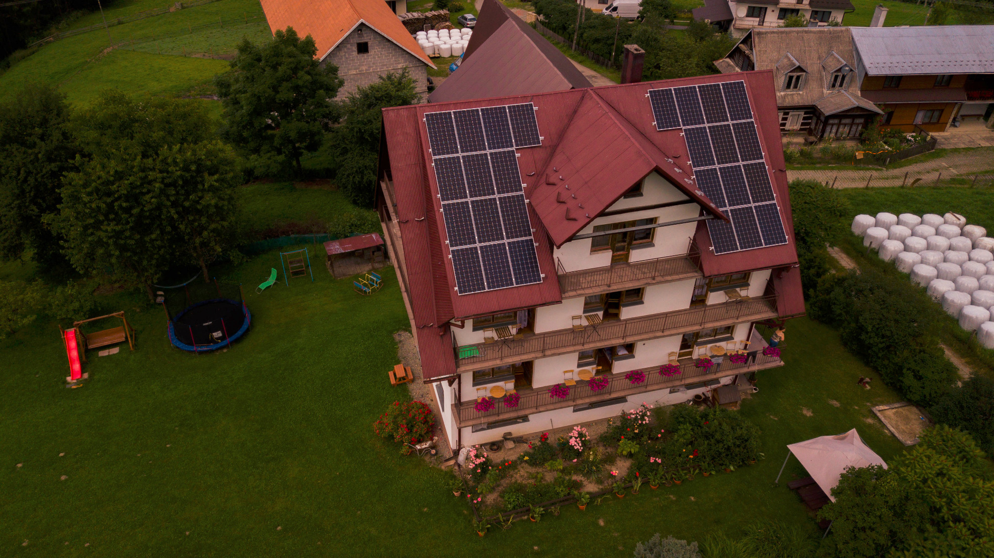 Widok z góry na dom z czerwonym dachem i panelami słonecznymi, otoczony zielonym trawnikiem z trampoliną i stogami siana w tle.