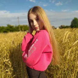 Portret młodej kobiety w różowej bluzie na tle pola pszenicy w słoneczny dzień. Dziewczyna ma długie blond włosy z różowymi pasemkami i delikatny makijaż.