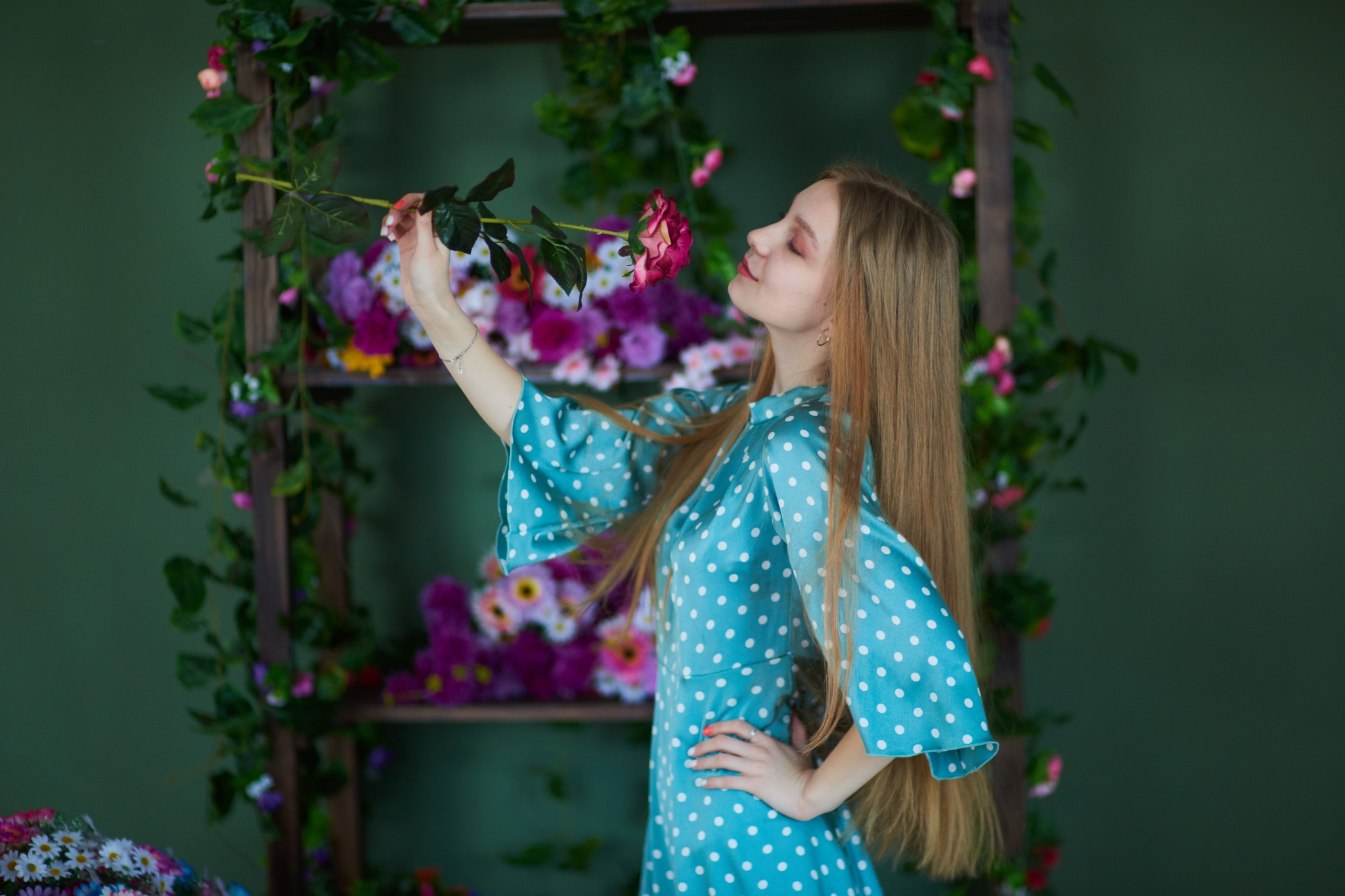 Młoda kobieta w błękitnej sukience w białe kropki wącha czerwoną różę na tle drewnianej konstrukcji z kwiatami.
