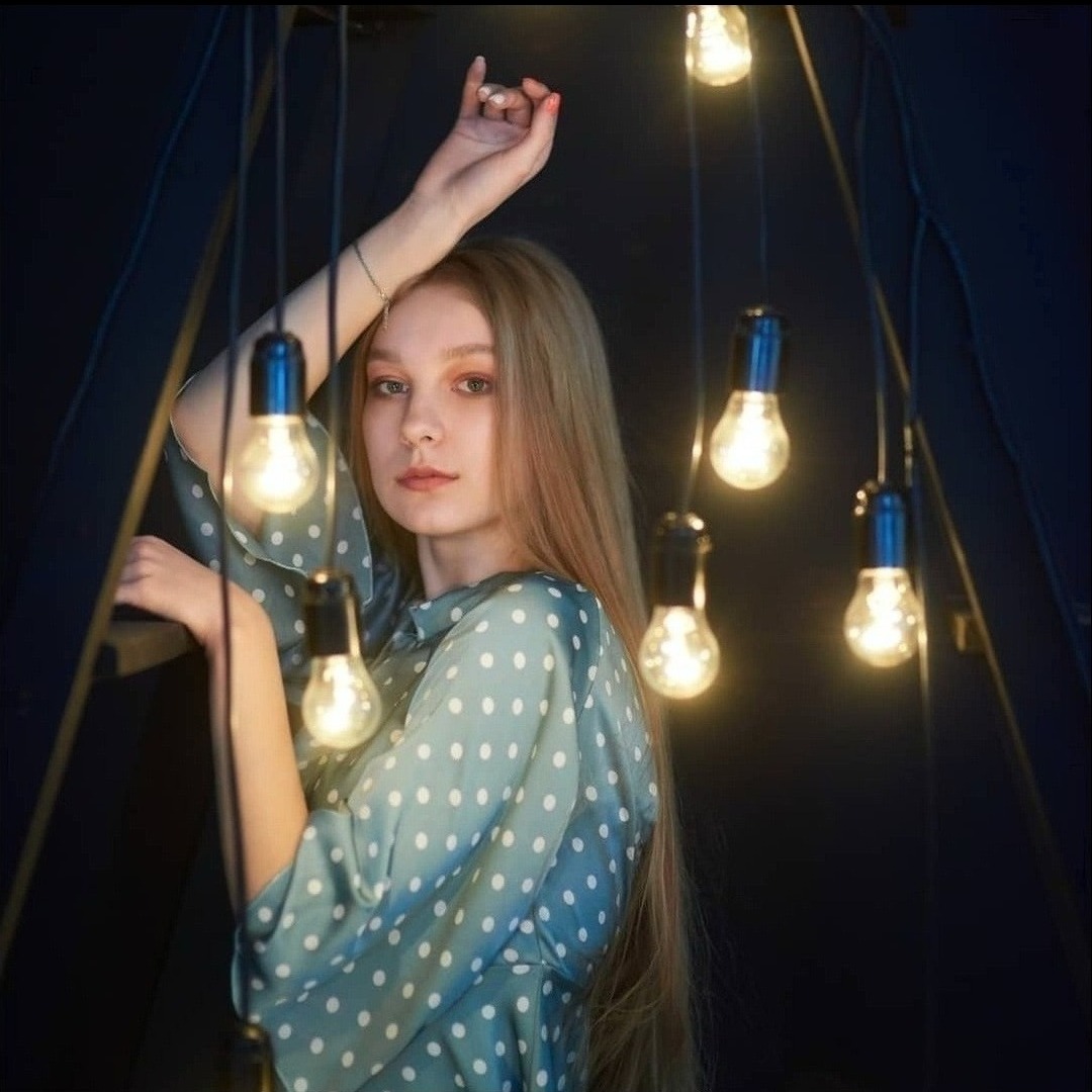 Portret kobiety w bluzce w kropki, otoczonej wiszącymi żarówkami na tle ciemnej ściany.