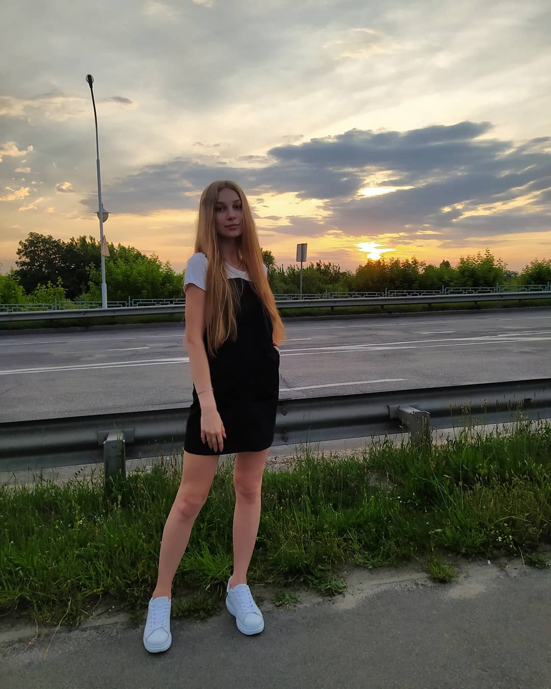 Młoda kobieta w czarnej sukience i białych butach pozuje na tle drogi i zachodzącego słońca.