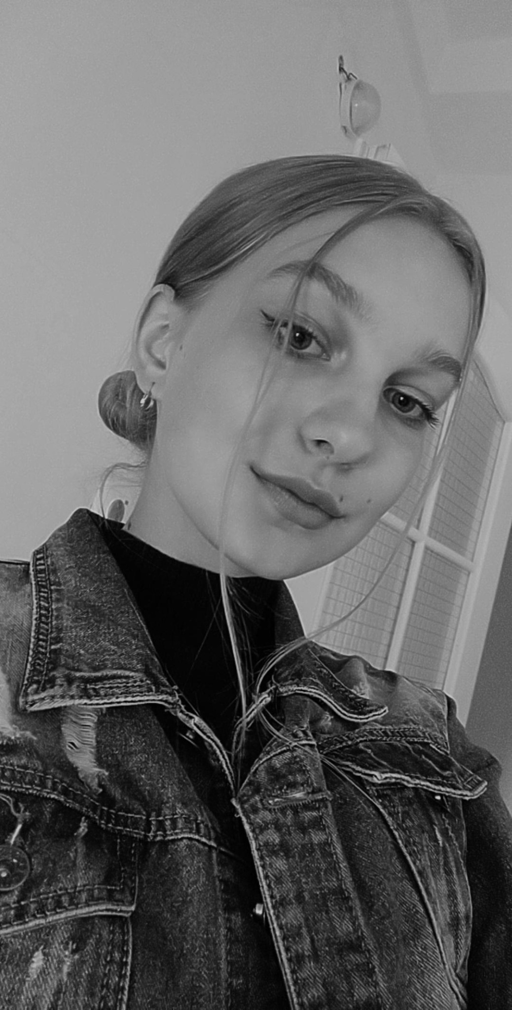 Portret młodej kobiety w dżinsowej kurtce na tle białych drzwi, zdjęcie czarno-białe.