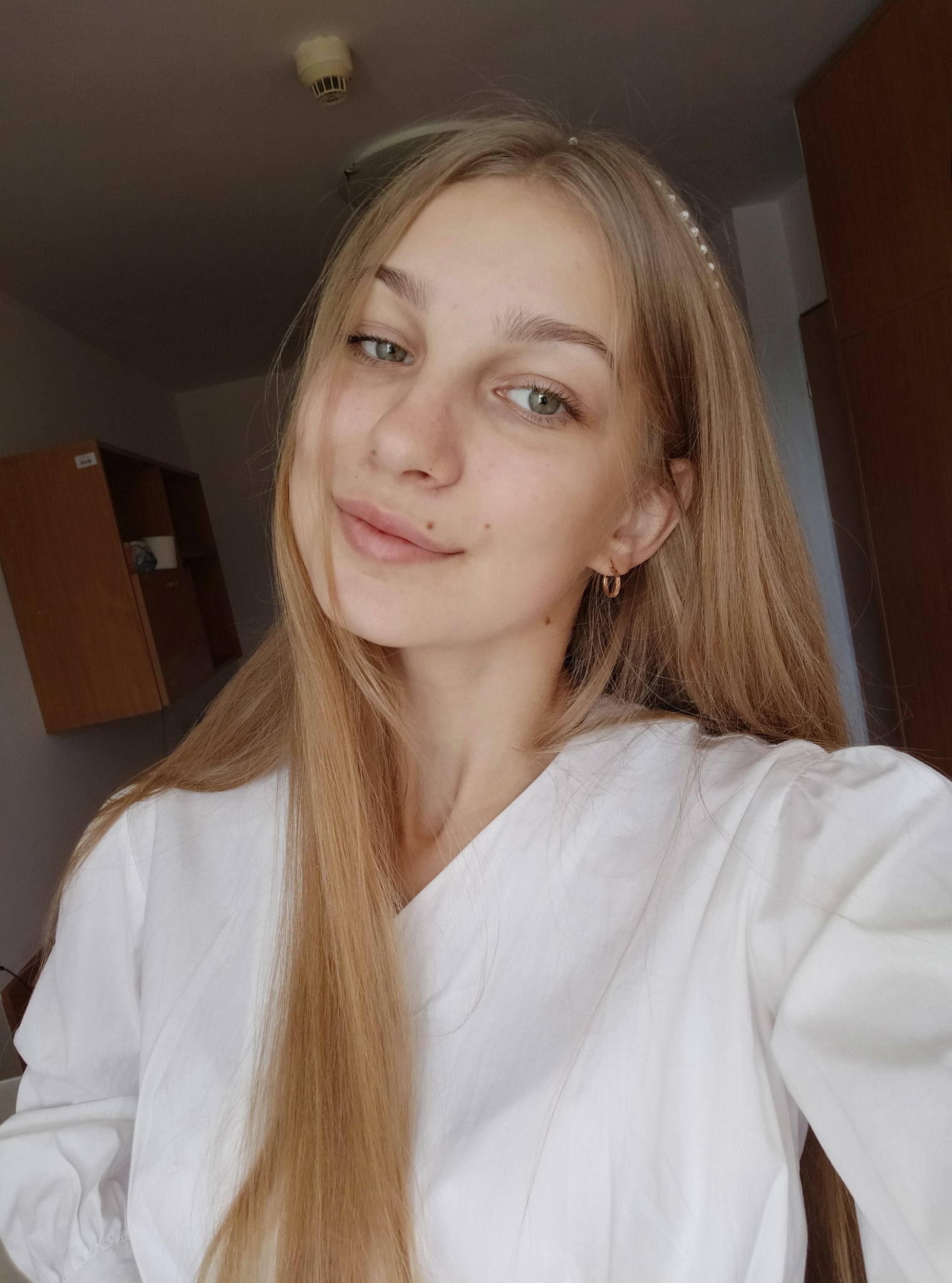 Portret uśmiechniętej blondynki w białej bluzce, z perłową opaską we włosach i złotymi kolczykami