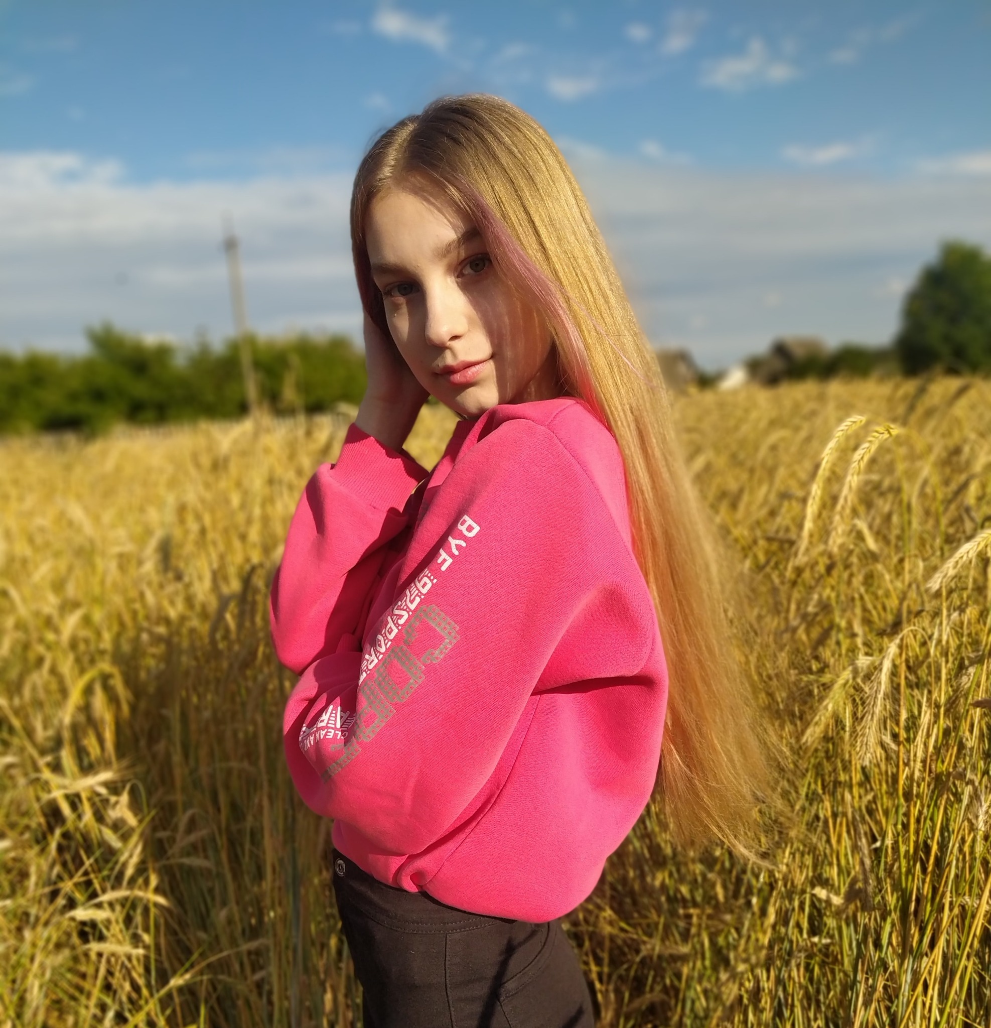 Portret młodej kobiety w różowej bluzie na tle pola pszenicy w słoneczny dzień. Dziewczyna ma długie blond włosy z różowymi pasemkami i delikatny makijaż.