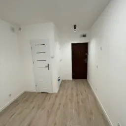 Wnętrze korytarza po remoncie: drewnopodobne panele podłogowe, białe ściany z listwami przypodłogowymi, białe drzwi wewnętrzne z poziomymi przeszkleniami, ciemne drzwi wejściowe, oświetlenie punktowe.