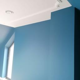 Damian Kowalski - Pomieszczenie po remoncie: błękitne ściany, biały sufit z listwami dekoracyjnymi i okrągłym nawiewnikiem wentylacyjnym, widoczne okno.