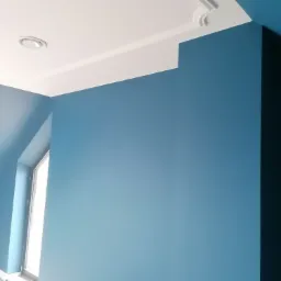 Pomieszczenie po remoncie: błękitne ściany, biały sufit z listwami dekoracyjnymi i okrągłym nawiewnikiem wentylacyjnym, widoczne okno.