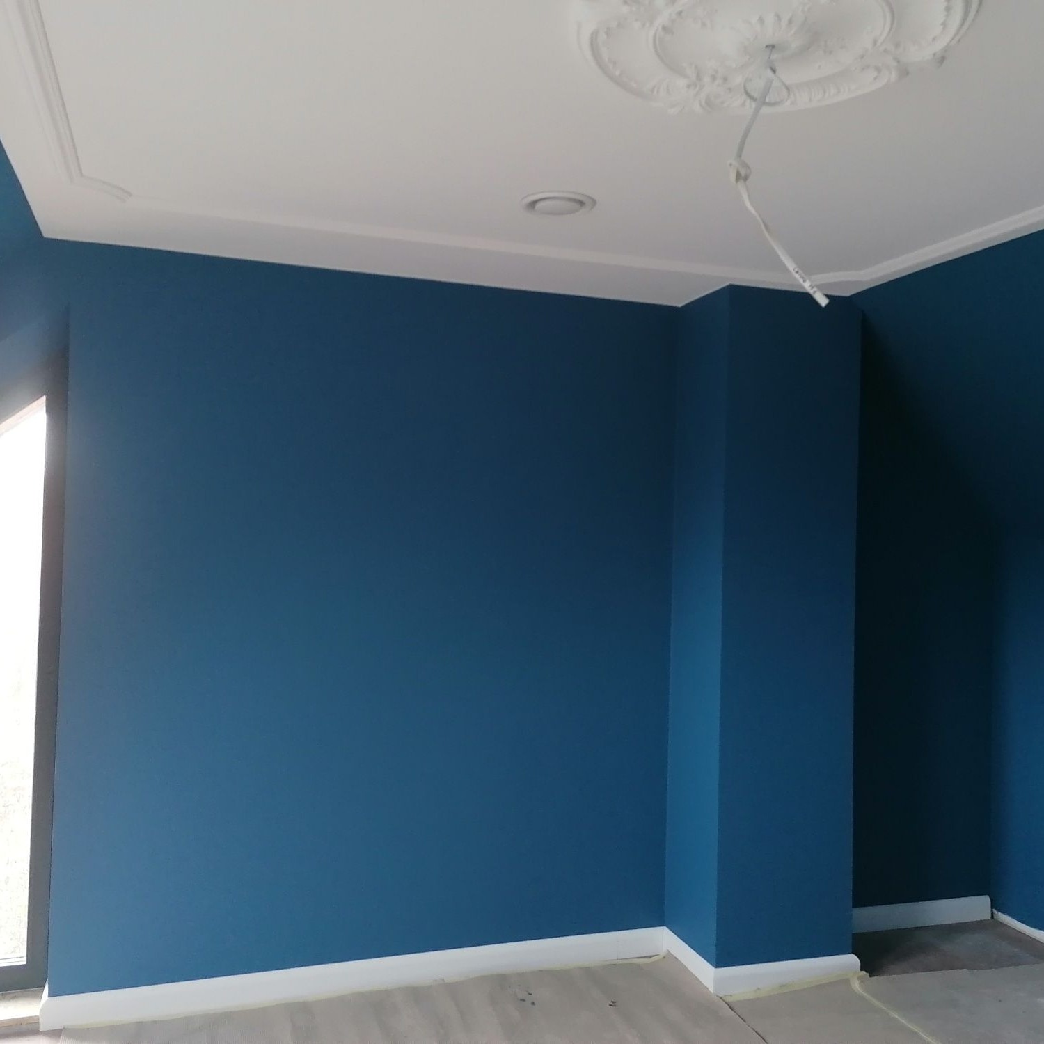 Świeżo pomalowane na niebiesko ściany w pokoju z białymi listwami przypodłogowymi i ozdobnym sztukateryjnym rozetą na suficie, widoczne kable wystające z sufitu.