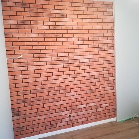 Ściana wykończona panelami imitującymi czerwoną cegłę z widocznym gniazdkiem elektrycznym, częściowo widoczna biała ściana po prawej stronie i fragment drewnopodobnej podłogi.