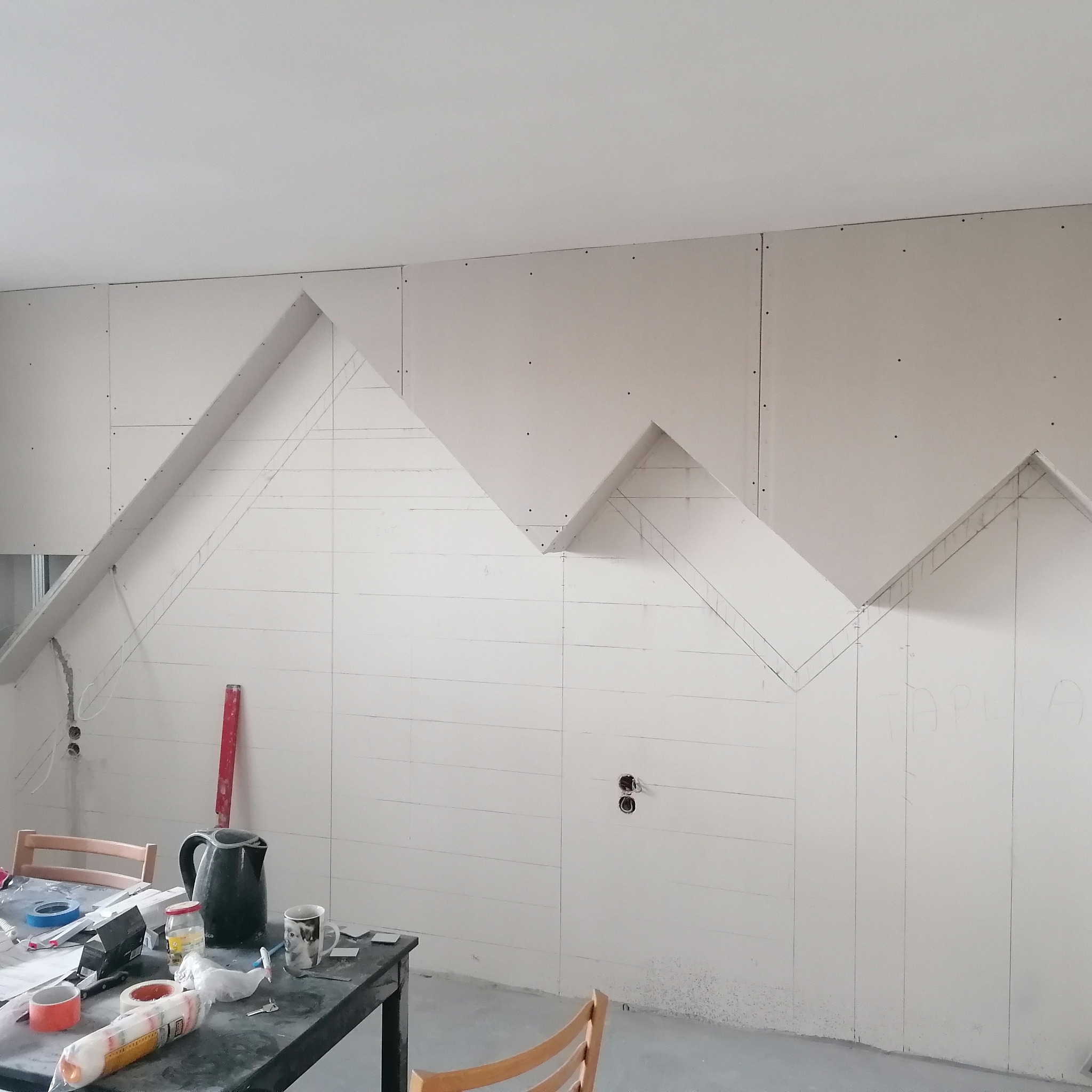 Ściana z płyt gipsowo-kartonowych w trakcie montażu, widoczne zaznaczone linie i kształty, stół z narzędziami i czajnikiem w tle, częściowo wykończone gniazdka elektryczne.