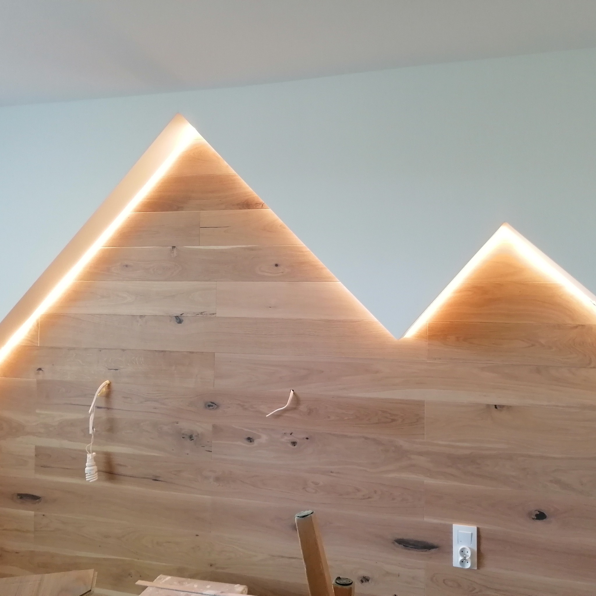 Drewniana ściana z podświetleniem LED w kształcie gór, widoczne wystające kable i gniazdko elektryczne.