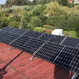 Panele słoneczne zamontowane na czerwonym, blaszanym dachu, z widokiem na zielone drzewa i zabudowania w tle.