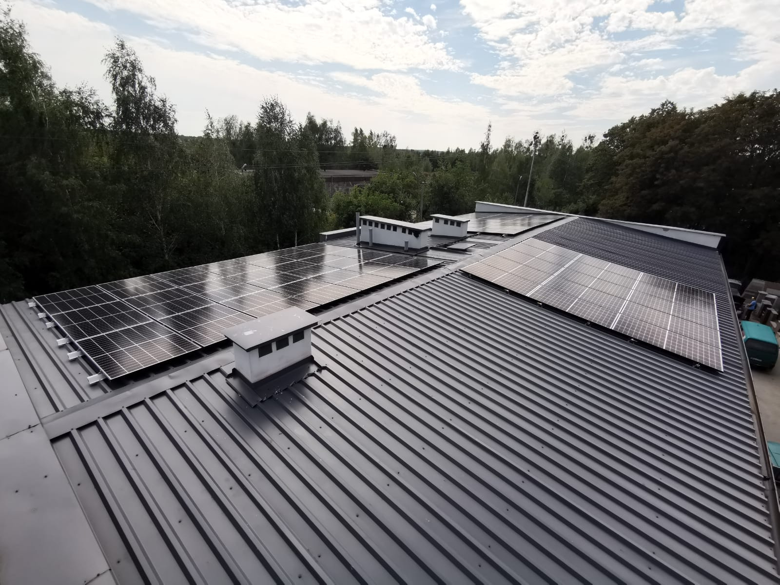 Instalacja paneli słonecznych na dachu z blachy trapezowej, widoczne kominki wentylacyjne, tło z drzewami i niebem.
