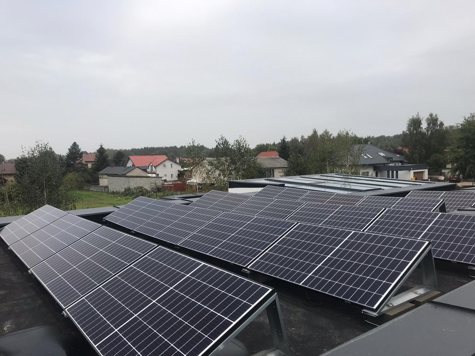 Rząd paneli słonecznych na płaskim dachu z widokiem na zabudowę jednorodzinną w tle podczas pochmurnego dnia.