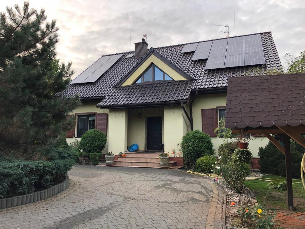 Dom jednorodzinny z brązowym dachem i panelami na dachu, widok z brukowanego podjazdu, dekoracje jesienne przed wejściem.