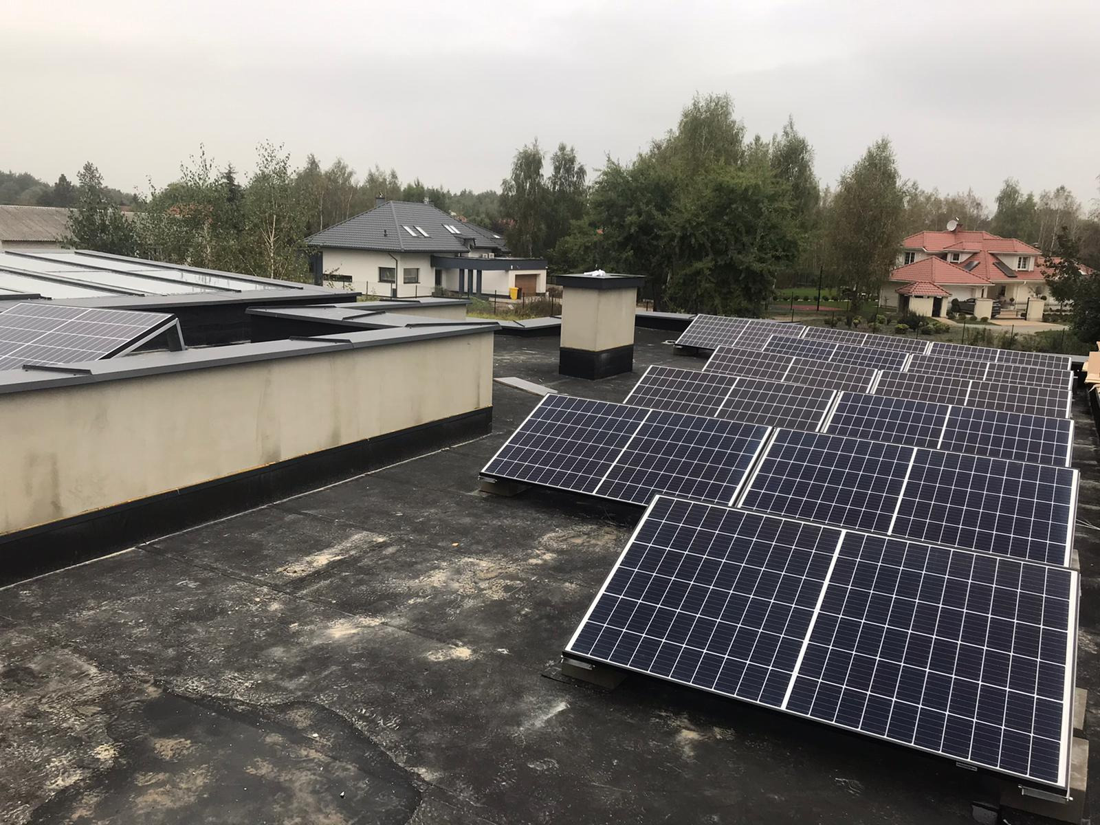 Rząd paneli słonecznych na płaskim dachu budynku z widokiem na domy jednorodzinne i drzewa w pochmurny dzień.