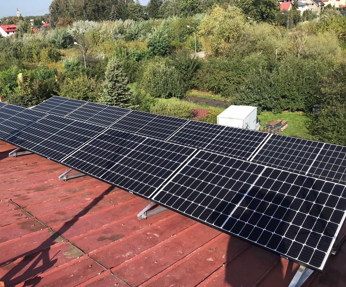 Panele słoneczne zamontowane na czerwonym, blaszanym dachu, z widokiem na zielone drzewa i zabudowania w tle.
