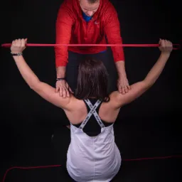 Kobieta siedzi na macie, rozciągając ramiona z czerwoną gumą pod nadzorem mężczyzny w czerwonej bluzie.