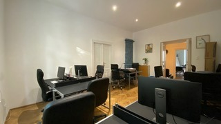 Przestronne biuro z parkietem, czarnymi biurkami i krzesłami obrotowymi, oświetlone naturalnym światłem, z widocznym fragmentem drugiego pomieszczenia w tle.