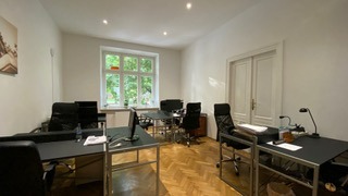 Przestronne biuro z trzema czarnymi biurkami i krzesłami, parkiet na podłodze, widok na zieleń za oknem, białe ściany i drzwi.