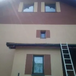 Dwukolorowa fasada domu z oknami w brązowych obramowaniach, z widoczną drabiną opartą o rynnę.