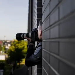 Mężczyzna w skórzanej kurtce z profesjonalnym aparatem fotograficznym z teleobiektywem, ukryty za murem z cegły, robi zdjęcia na tle zabudowań we Wrocławiu.