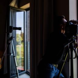 Detektyw i Bezpieczeństwo OPERAND - Mężczyzna w ciemnym pomieszczeniu, przy oknie, filmuje krajobraz za pomocą profesjonalnej kamery na statywie; drugi statyw z kamerą widoczny w tle.
