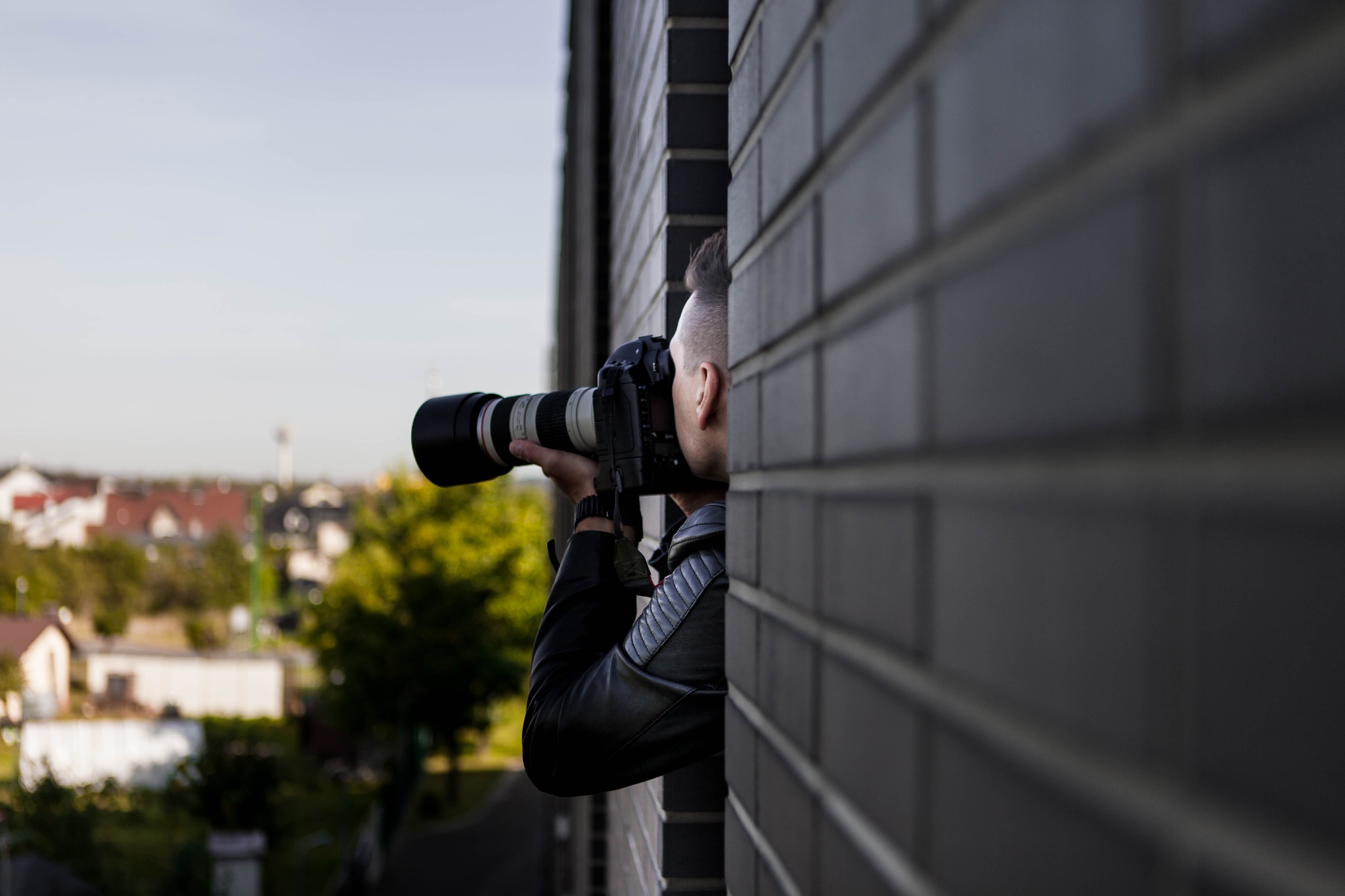 Mężczyzna w skórzanej kurtce z profesjonalnym aparatem fotograficznym z teleobiektywem, ukryty za murem z cegły, robi zdjęcia na tle zabudowań we Wrocławiu.