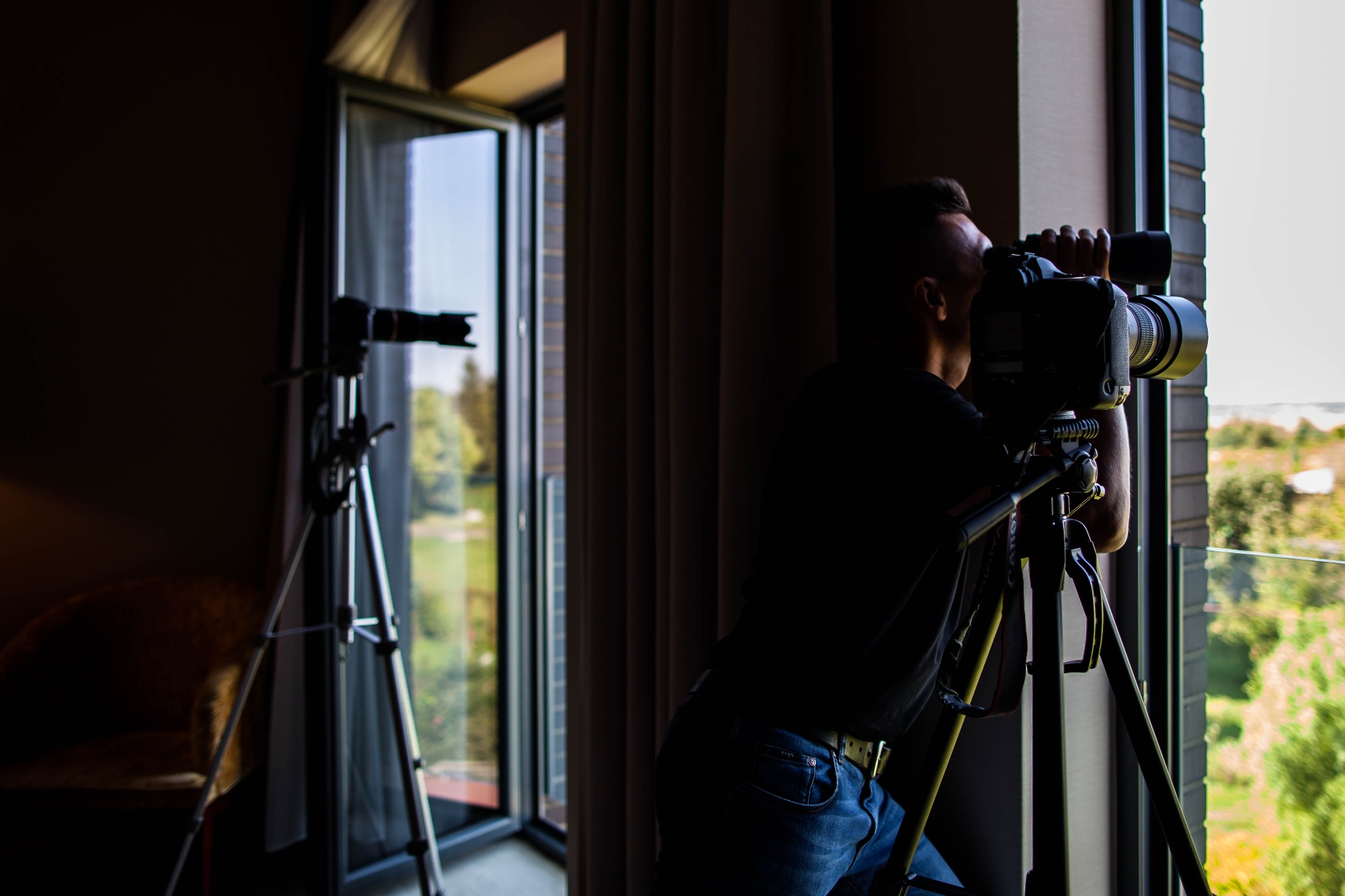 Mężczyzna w ciemnym pomieszczeniu, przy oknie, filmuje krajobraz za pomocą profesjonalnej kamery na statywie; drugi statyw z kamerą widoczny w tle.