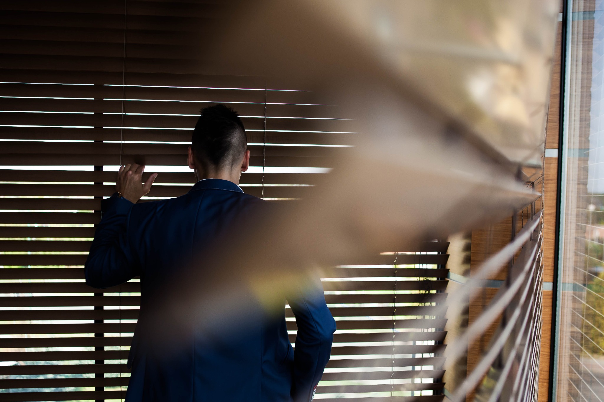 Mężczyzna w granatowym garniturze dyskretnie spogląda przez drewniane żaluzje, z widocznym zegarkiem na nadgarstku, częściowo zasłonięty przez rozmyte elementy żaluzji na pierwszym planie.