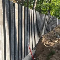 Nowoczesne ogrodzenie z prefabrykowanych, pionowych elementów betonowych w trakcie montażu, widoczna niwelacja terenu i drabina oparta o konstrukcję.