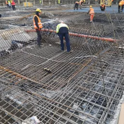 Siatka zbrojeniowa ułożona na placu budowy, pracownicy w kamizelkach odblaskowych i kaskach montują konstrukcję, widoczne rury i przewody.