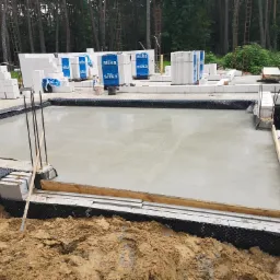 Świeżo wylana wylewka fundamentowa z widocznymi wystającymi prętami zbrojeniowymi, bloczkami Silka i hydroizolacją w otoczeniu ziemi i drzew.