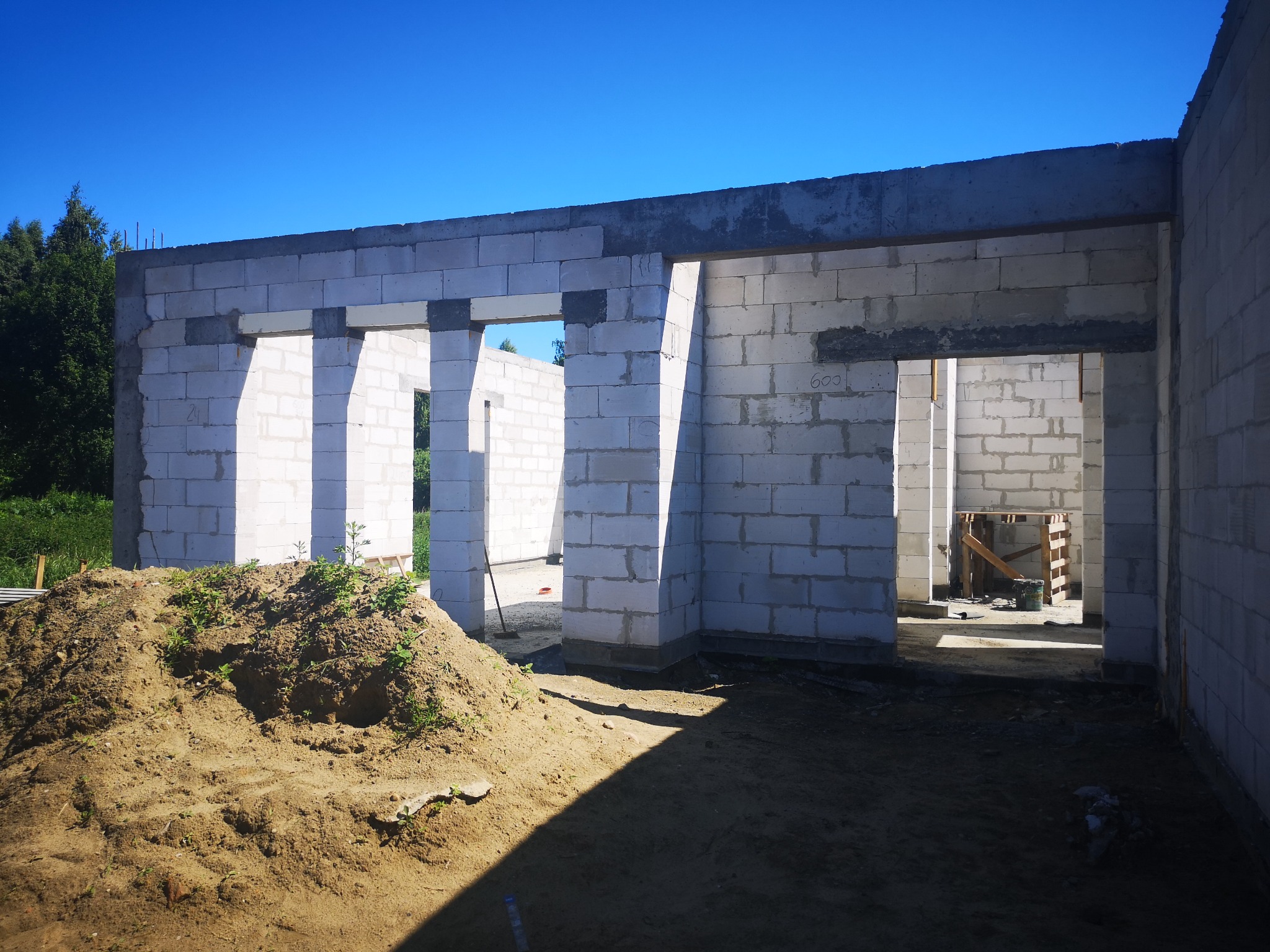 Surowy stan budowy domu z białych bloczków, widoczny stos ziemi przed budynkiem i błękitne niebo w tle.